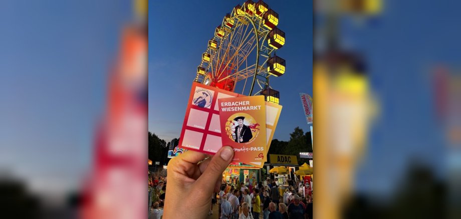 Wiesemaiks-Pass 2026 Karte mit Aufschrift "Wiesemaiks-Pass" wird von Hand hochgehalten. Im Hintergrund ist ein Riesenrad bei Nacht zu sehen.