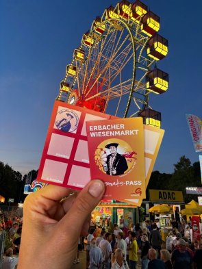 Karte mit Aufschrift "Wiesemaiks-Pass" wird von Hand hochgehalten. Im Hintergrund ist ein Riesenrad bei Nacht zu sehen.