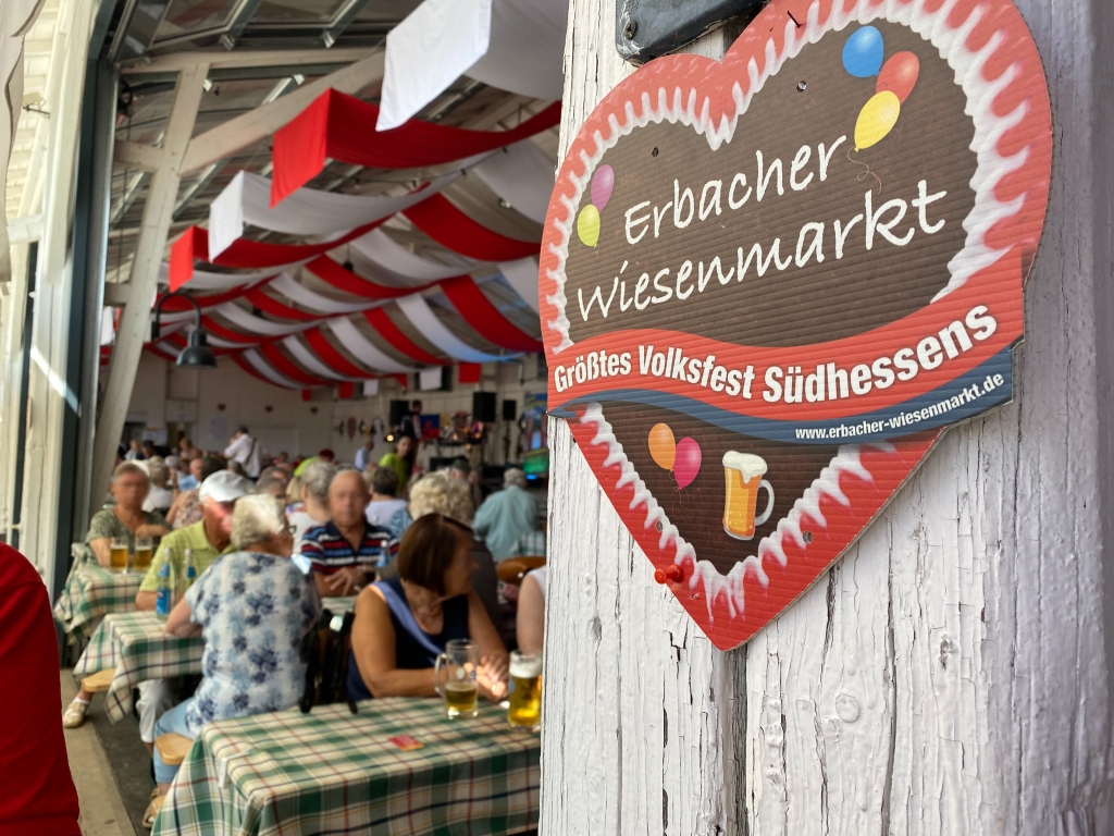 Geschmückte Festhalle, in der ältere Menschen mit Bier und anderen Getränken an Biertischgarnituren sitzen. Im Vordergrund rechts ist ein Herz mit der Aufschrift Erbacher Wiesenmarkt zu sehen.