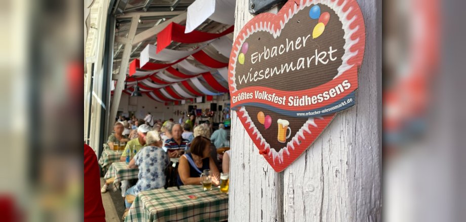 Seniorenfrüschoppen Geschmückte Festhalle, in der ältere Menschen mit Bier und anderen Getränken an Biertischgarnituren sitzen. Im Vordergrund rechts ist ein Herz mit der Aufschrift Erbacher Wiesenmarkt zu sehen.