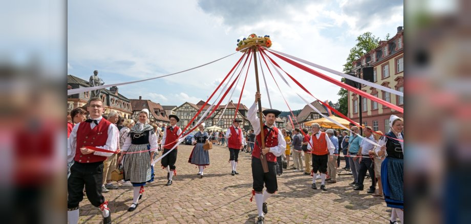 Menschen in traditioneller Tracht wirken bei Umzug mit und halten Baum mit Bändern hoch.