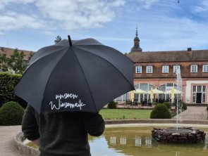 Schwarzer aufgeklappter Schirm mit Schriftzug "unsern Wiesemaik" vor Parkkulisse