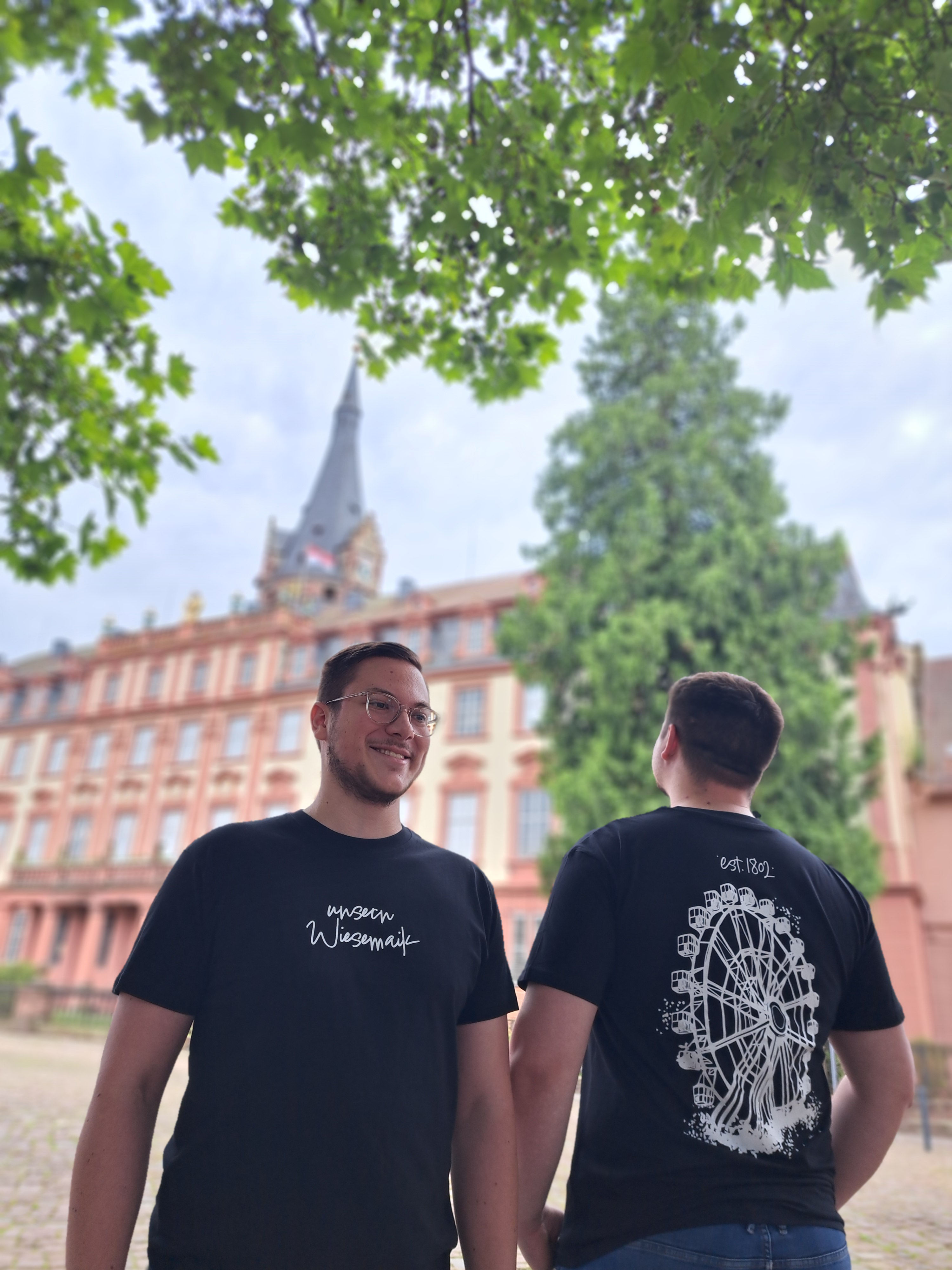 Zwei Männer stehen vor einem Schloss und tragen je ein schwarzes T-Shirt. Auf den T-Shirts ist einmal ein Riesenrad und einmal der Schriftzug "unsern Wiesemaik" zu sehen.