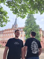 Zwei Männer stehen vor einem Schloss und tragen je ein schwarzes T-Shirt. Auf den T-Shirts ist einmal ein Riesenrad und einmal der Schriftzug "unsern Wiesemaik" zu sehen.