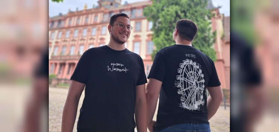 T-Shirt "unsern Wiesemaik" Zwei Männer stehen vor einem Schloss und tragen je ein schwarzes T-Shirt. Auf den T-Shirts ist einmal ein Riesenrad und einmal der Schriftzug "unsern Wiesemaik" zu sehen.