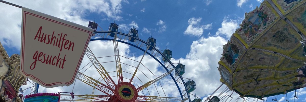Rummelplatz mit Riesenrad und Kettenflieger. Im Vordergrund ist ein Schild mit der Aufschrift Aushilfen gesucht zu sehen