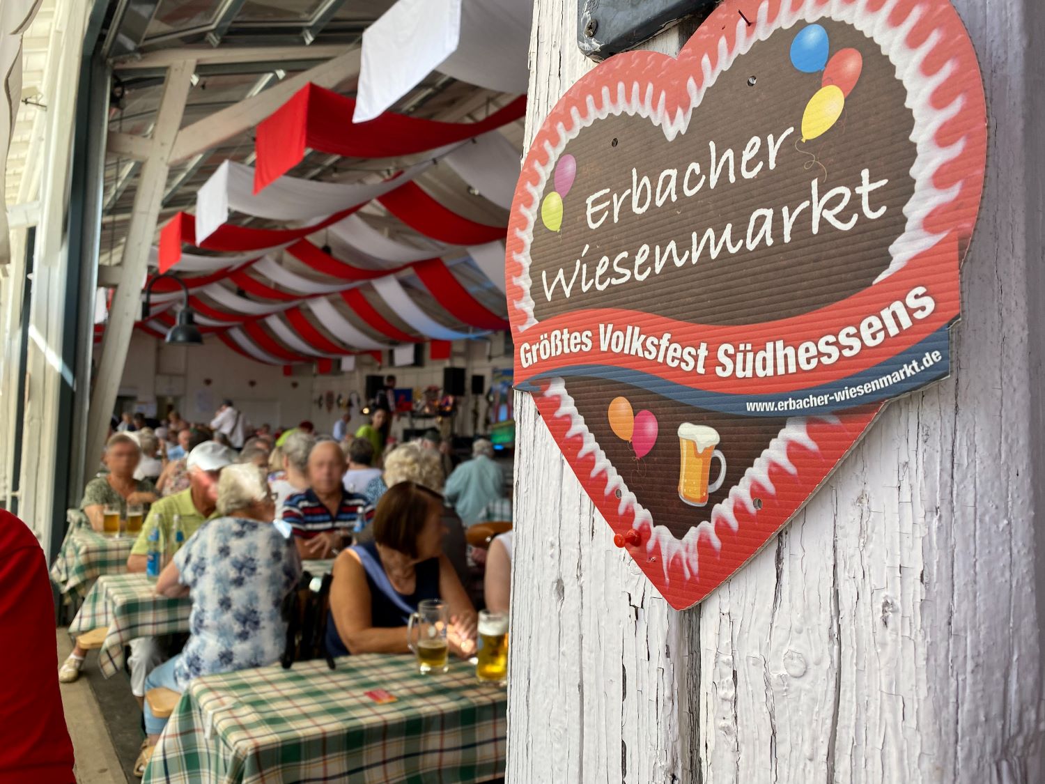 Großes Pappherz mit Aufschrift "Erbacher Wiesenmarkt" im Vordergrund, Menschen an Biertischgarnituren im Hintergrund