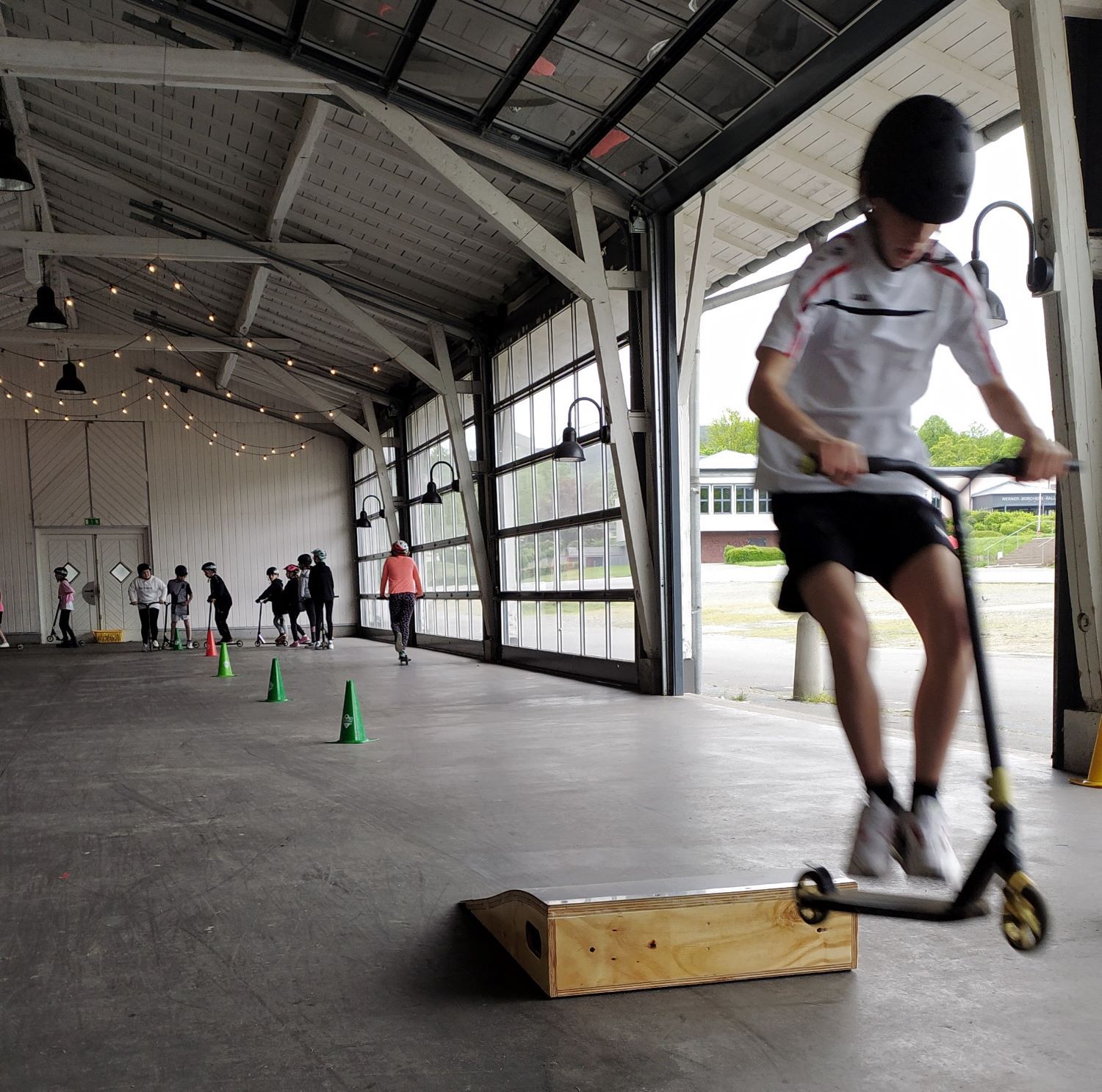 Kind springt mit Scooter über Hindernis. Im Hintergrund sind weitere Kinder beim Scooter-Fahren zu sehen.