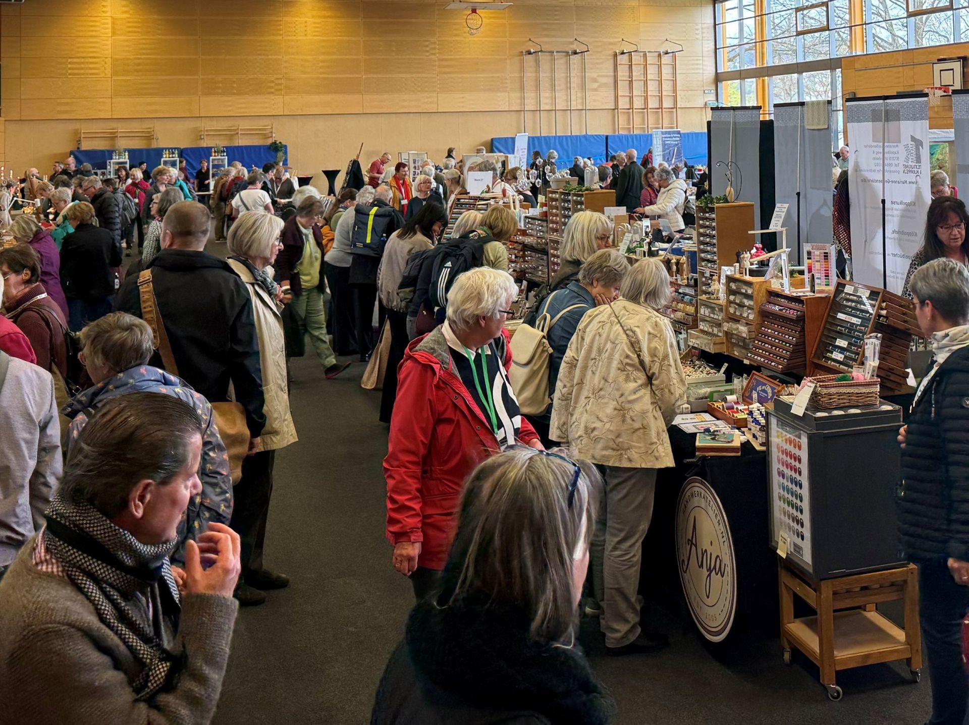 Viele Personen, die sich Waren (Spitzendecken) von Händlern in einer Sporthalle ansehen.
