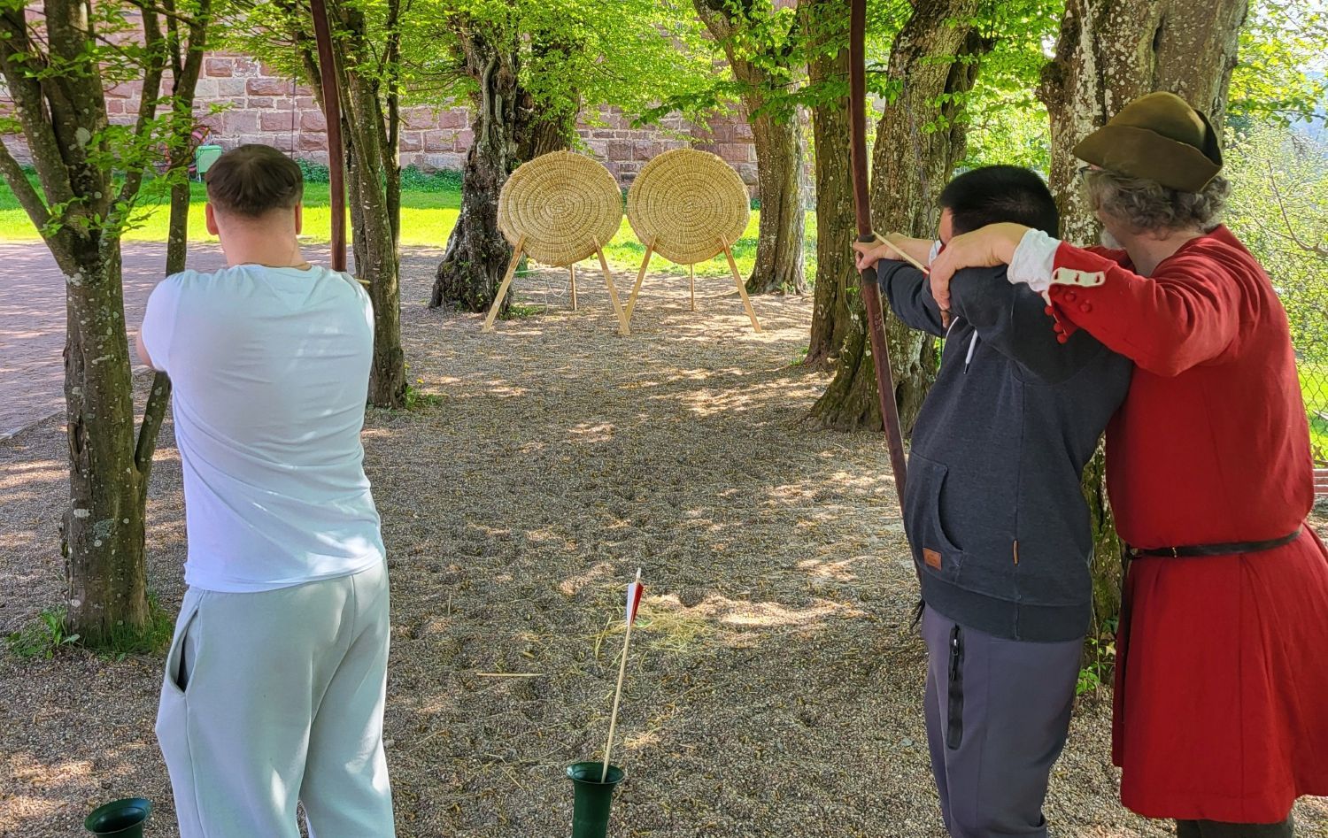 Zwei Jugendliche beim Bogenschießen auf eine Zielscheibe. Ein Mann in Mittelaltertracht leitet sie dabei an.