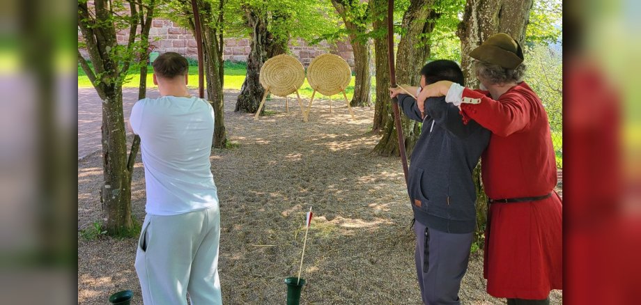 Zwei Jugendliche beim Bogenschießen auf eine Zielscheibe. Ein Mann in Mittelaltertracht leitet sie dabei an.