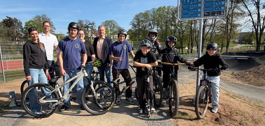 Erwachsene und Kinder stehen auf Pumptrack-Platz. Kinder mit Helm und Fahrrad.