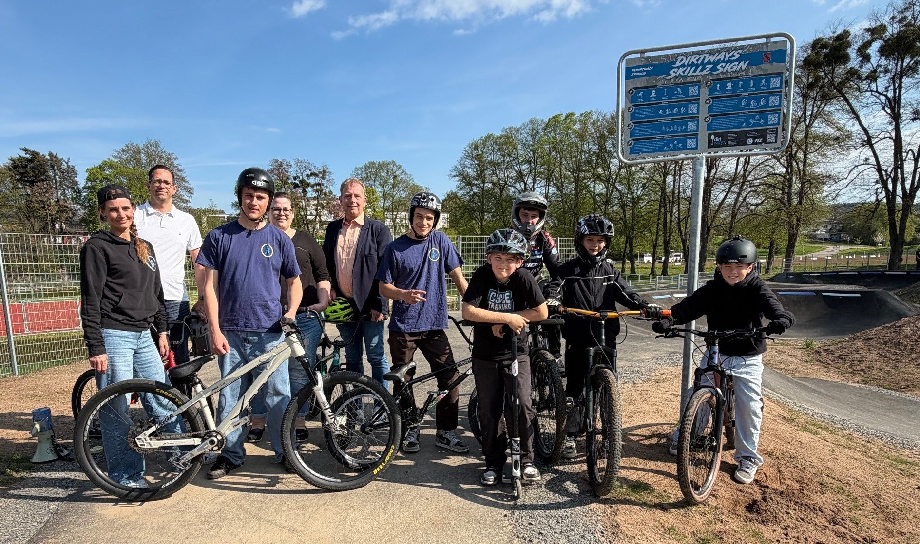 Erwachsene und Kinder stehen auf Pumptrack-Platz. Kinder mit Helm und Fahrrad.