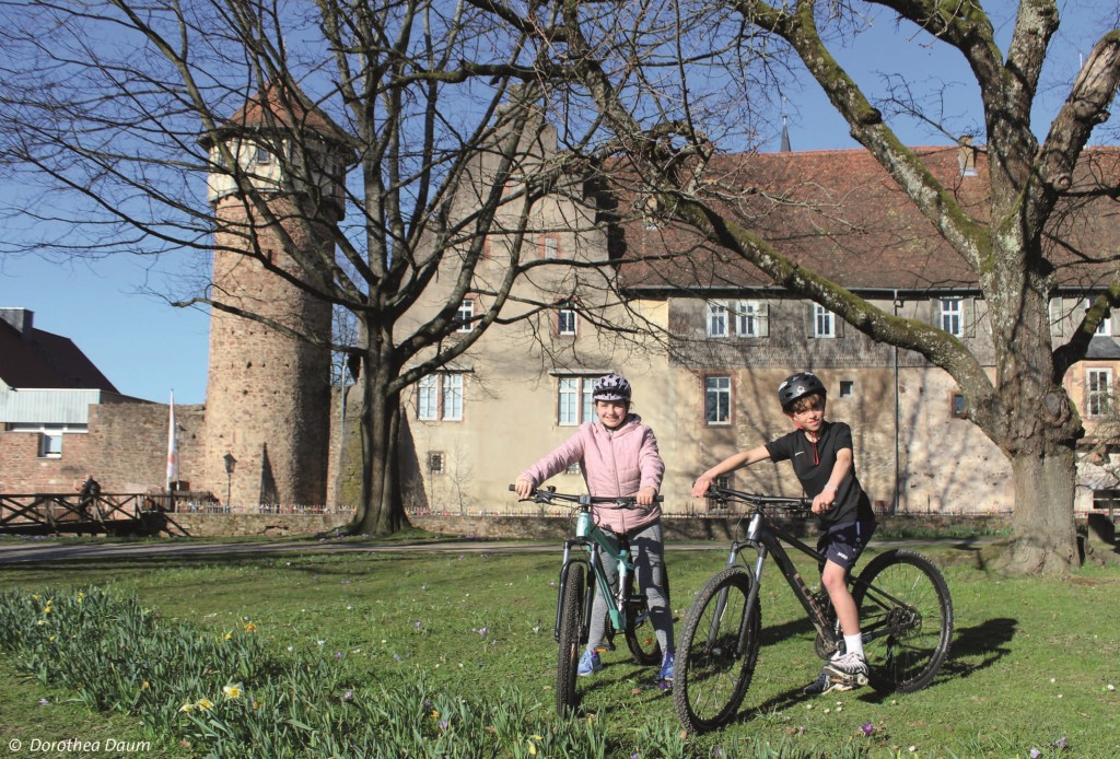 Zwei Kinder auf Fahrrädern lächeln vor einem historischen Gebäude in die Kamera