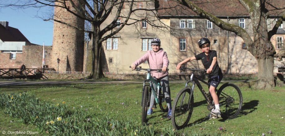 Zwei Kinder auf Fahrrädern lächeln vor einem historischen Gebäude in die Kamera