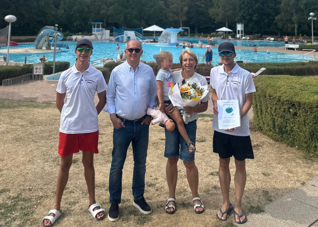 Eine Frau mit kleinem Kind auf dem Arm hält einen Blumenstrauß in den Händen. Drei Männer stehen daneben. Im Hintergrund ist ein Freibad zu sehen.