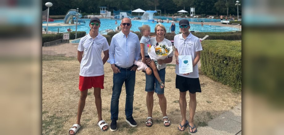 Marion Kaisers ist der 50.000. Badegast der Saison 2026 im Alexanderbad Eine Frau mit kleinem Kind auf dem Arm hält einen Blumenstrauß in den Händen. Drei Männer stehen daneben. Im Hintergrund ist ein Freibad zu sehen.