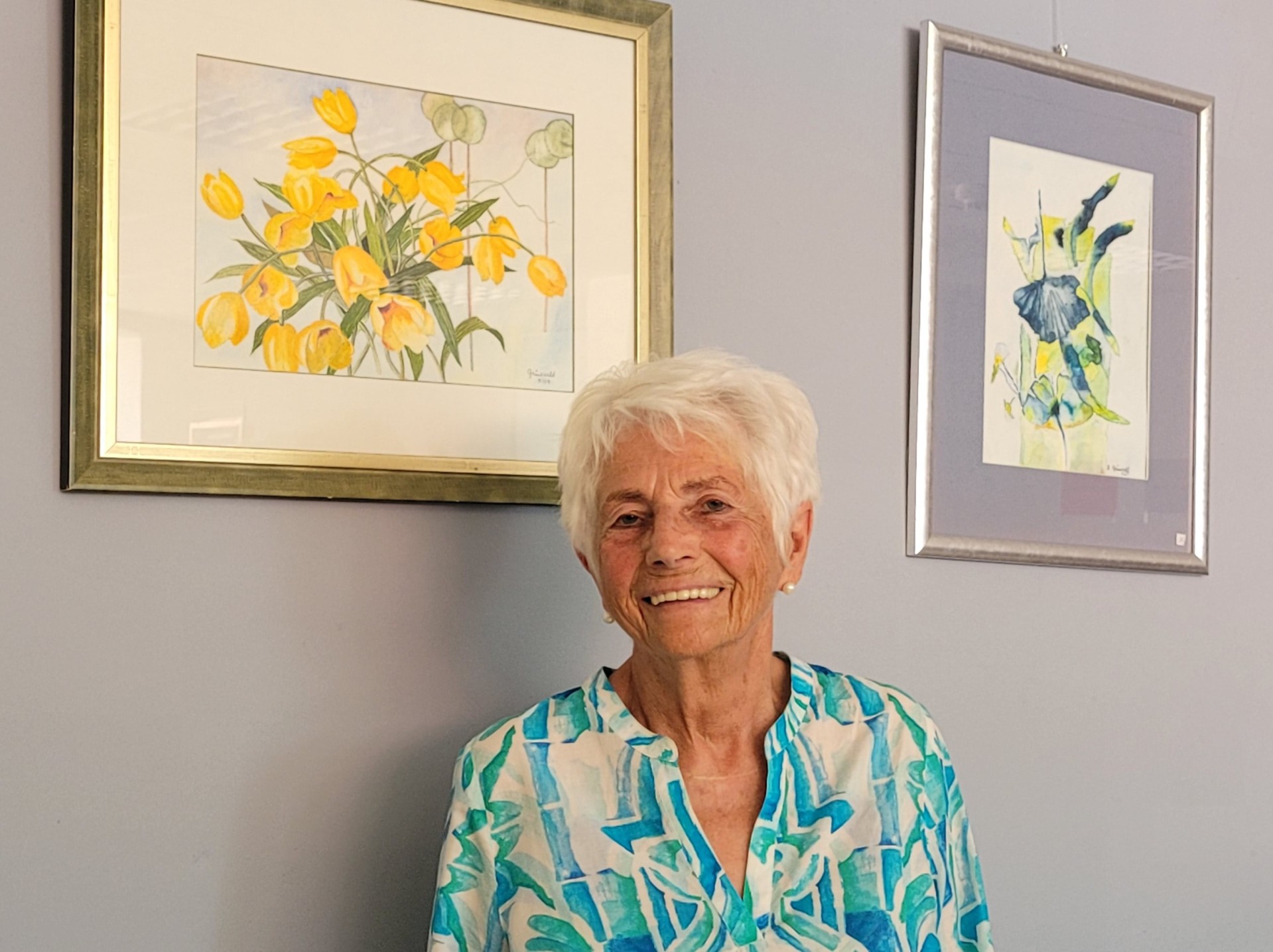 Eine Frau steht vor zwei Bilder mit Blumenmotiv und lächelt in die Kamera.