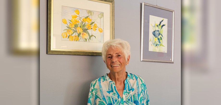 Eine Frau steht vor zwei Bilder mit Blumenmotiv und lächelt in die Kamera.