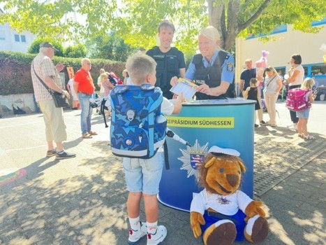 Eine Landespolizistin und ein Stadtpolizist stehen an einem Informationsstand vor einer Schule und überreichen einem Erstklässler einen Stundenplan.