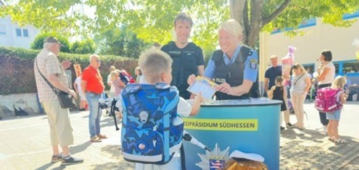 Informationsstand der Polizei für Grundschüler Eine Landespolizistin und ein Stadtpolizist stehen an einem Informationsstand vor einer Schule und überreichen einem Erstklässler einen Stundenplan.