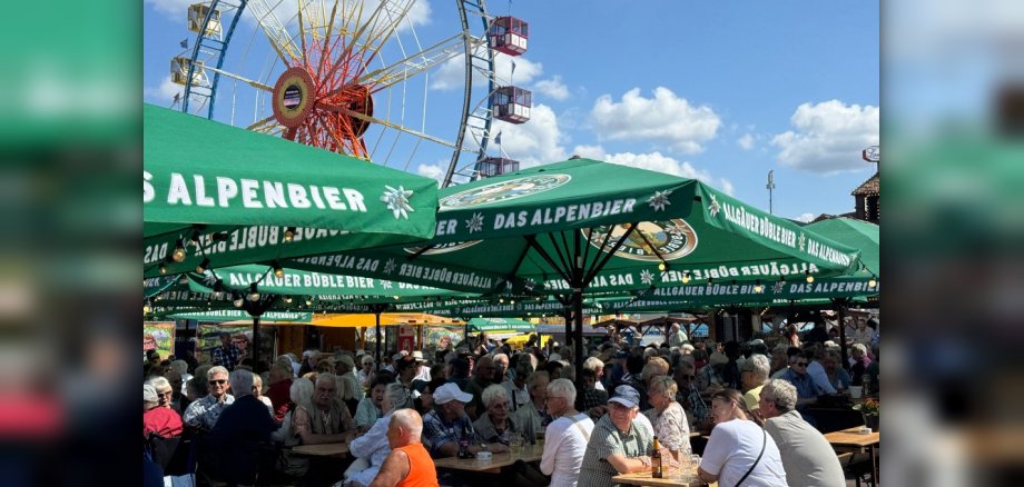 Seniorenfrühschoppen 2026 Seniorinnen und Senioren sitzen auf Biertischgarnituren in einem Biergarten. Im Hintergrund ist ein Riesenrad zu sehen.