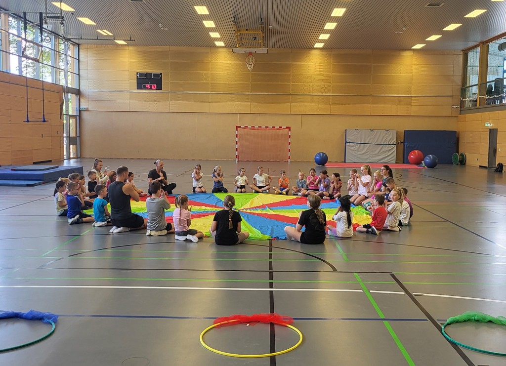 Kinder sitzen in einer Turnhalle im Kreis. In der Hand halten sie ein buntes großes Ballontuch.