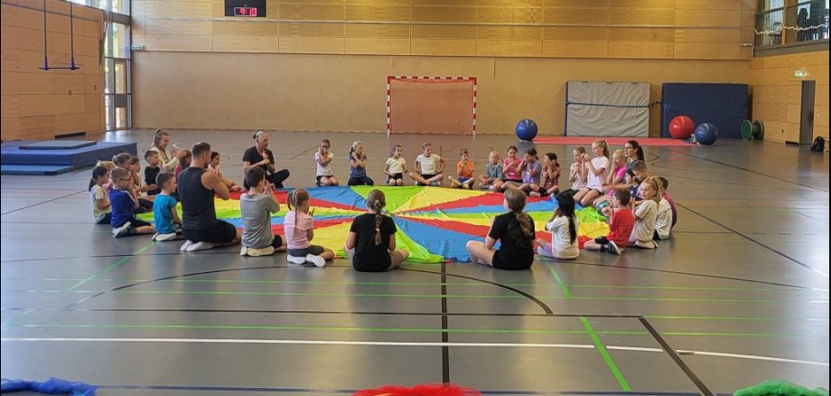 Kinder sitzen in einer Turnhalle im Kreis. In der Hand halten sie ein buntes großes Ballontuch.