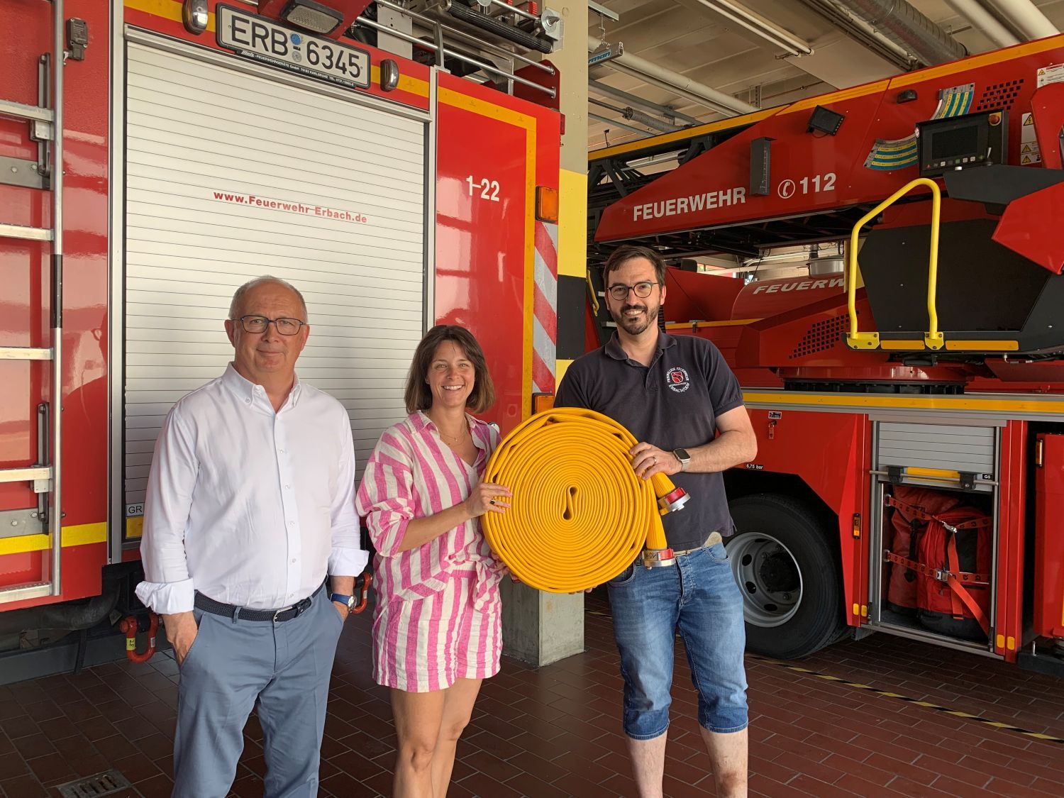 Zwei Männer und eine Frau stehen in einem Feuerwehrhaus vor einem Feuerwehrfahrzeug und halten einen zusammengerollten Feuerwehrschlauch in der Hand.