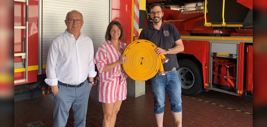 Übergabe der Feuerwehrschläuche Zwei Männer und eine Frau stehen in einem Feuerwehrhaus vor einem Feuerwehrfahrzeug und halten einen zusammengerollten Feuerwehrschlauch in der Hand.