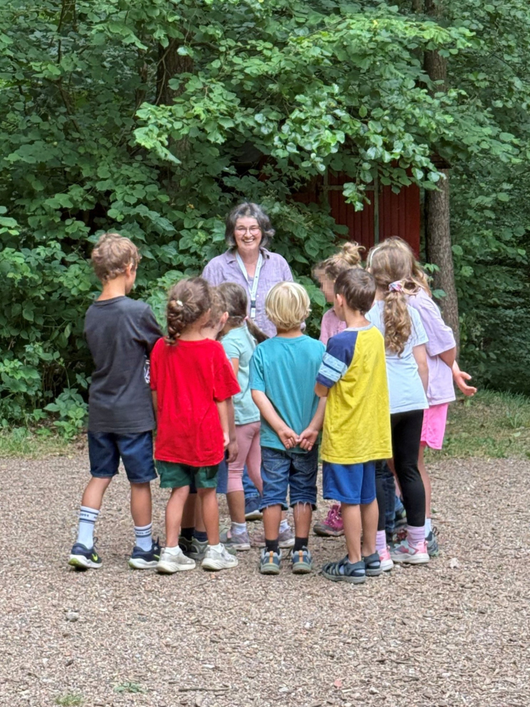 Eine Gruppe von Kindern steht im Wald vor einer Frau, die etwas erzählt. 