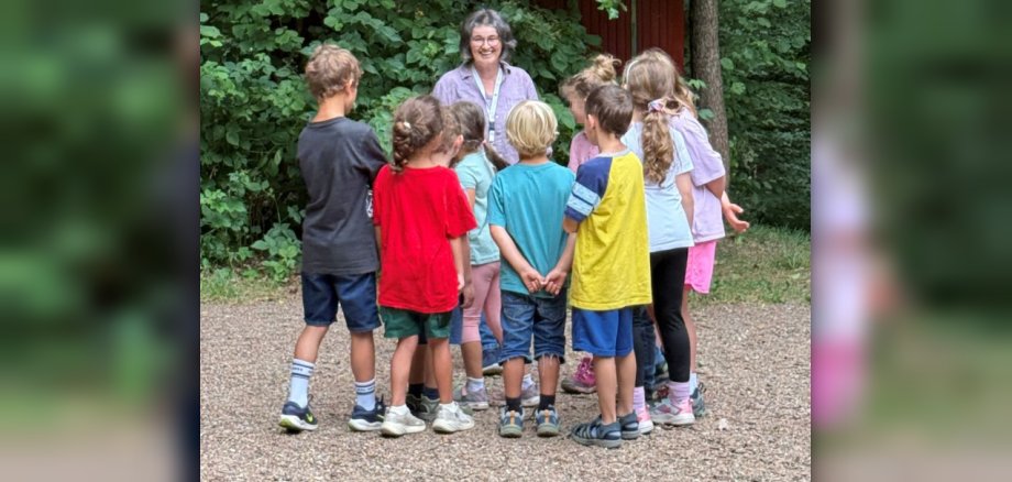 Eine Gruppe von Kindern steht im Wald vor einer Frau, die etwas erzählt. 