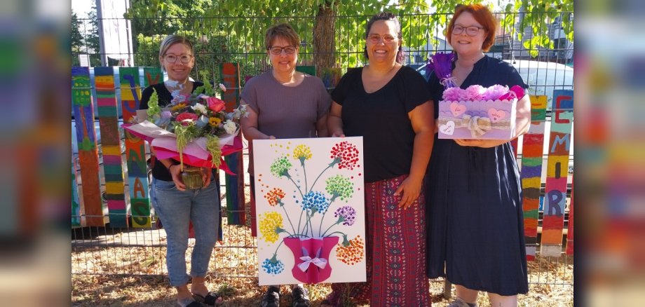 Vier Frauen blicken lächelnd in die Kamera. Zwei von ihnen halten je einen Blumenstrauß und einen Präsentkorb in den Händen, die anderen beiden halten ein Bild mit Blumen darauf fest.
