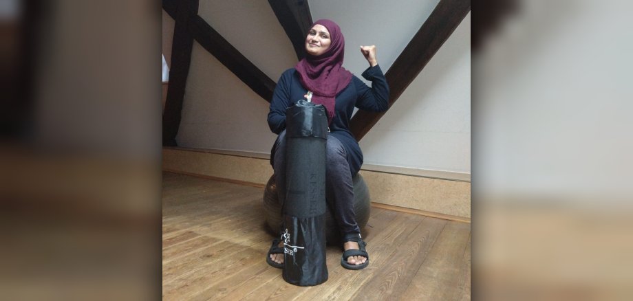 Frau mit Hijab hält eine Yoga-Matte in der Hand und hat den Arm gebeugt als Zeichen von Stärke