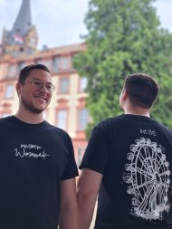 Zwei Männer stehen vor einem Schloss und tragen je ein schwarzes T-Shirt. Auf den T-Shirts ist einmal ein Riesenrad und einmal der Schriftzug "unsern Wiesemaik" zu sehen.