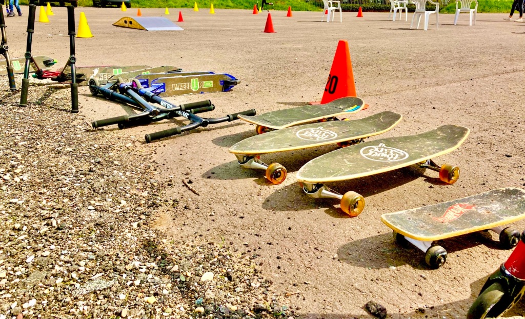 Skateboards und Scooter liegen auf einem Sandplatz. Dahinter sind Pylonen und Stühle zu sehen.