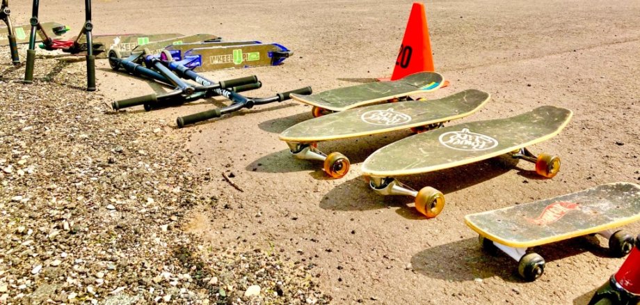 Spielfest für Kinder Skateboards und Scooter liegen auf einem Sandplatz. Dahinter sind Pylonen und Stühle zu sehen.