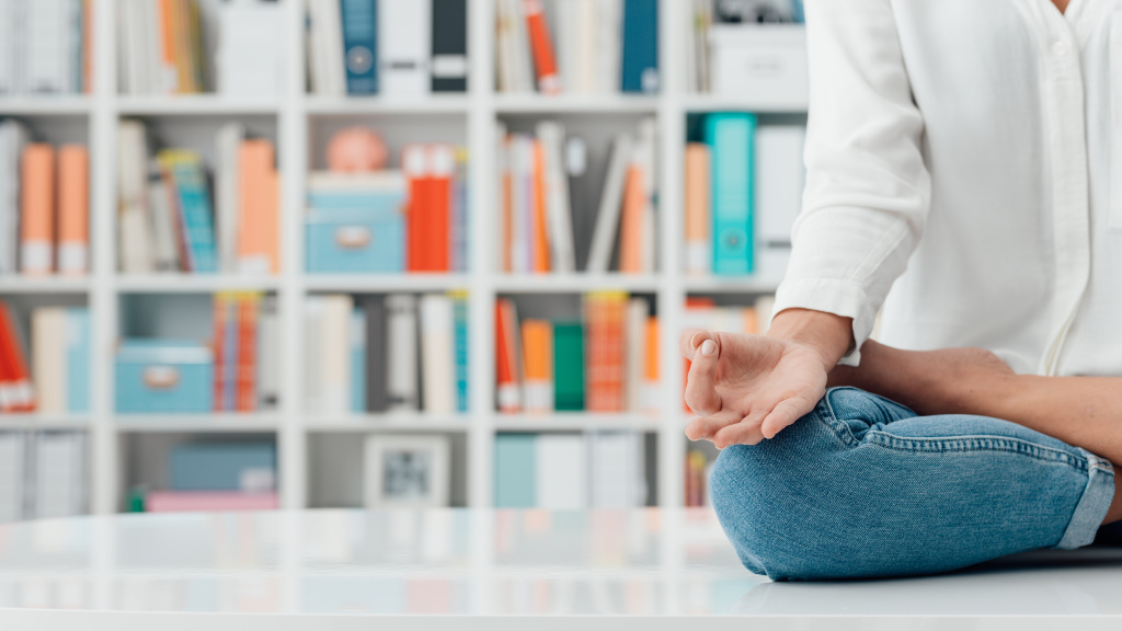 Eine Frau sitzt vor einer Bücherwand und meditiert im Schneidersitz