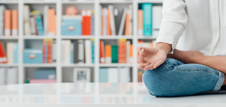 Achtsamkeit Eine Frau sitzt vor einer Bücherwand und meditiert im Schneidersitz