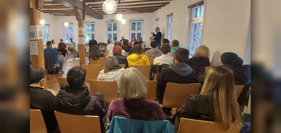 Bild einer Diskussionsrunde in einem Raum. Ein Mann steht mit Mikrofon in der Hand vor Publikum, das auf Stühlen sitzt.
