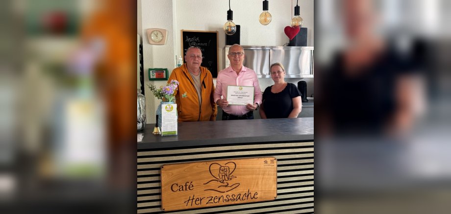 Übergabe der Patenschaftsurkunde Drei Menschen - zwei Männer und eine Frau - stehen hinter einem Tresen. Der Mann in der Mitte hält eine Urkunde hoch.