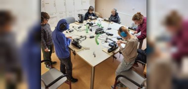 Schnitzen mit Naturmaterialien Kinder stehen an einem Tisch und tragen Ohrenschützer. Vor ihnen stehen Schneide- und Fräsmaschinen auf dem Tisch.