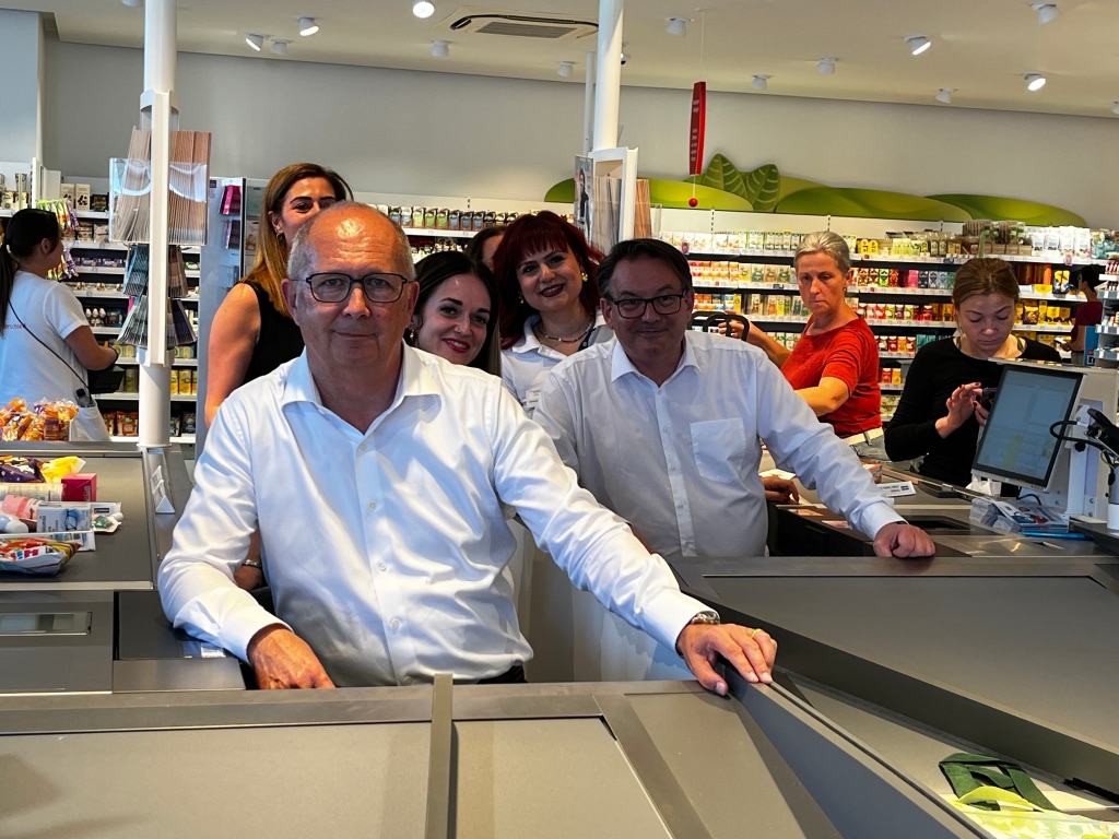 Zwei Männer sitzen am Kassierband bei dm. Dahinter Mitarbeiterinnen und Kunden