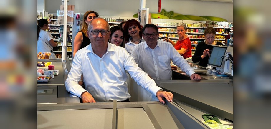 Zwei Männer sitzen am Kassierband bei dm. Dahinter Mitarbeiterinnen und Kunden