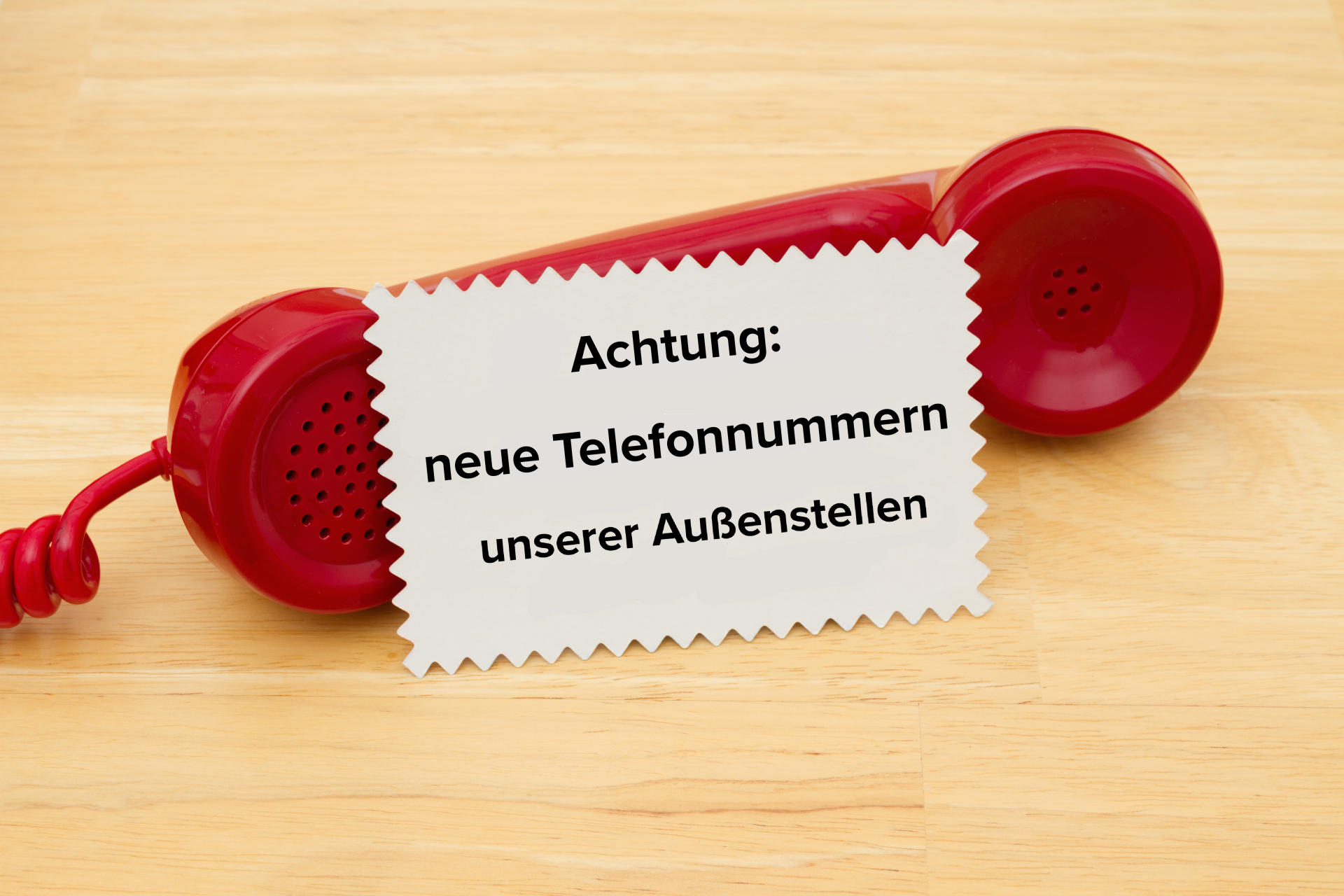 Roter Telefonhörer mit Telefonschnur. Davor ein weißes Kärtchen mit Hinweistext zu neuen Telefonnummern