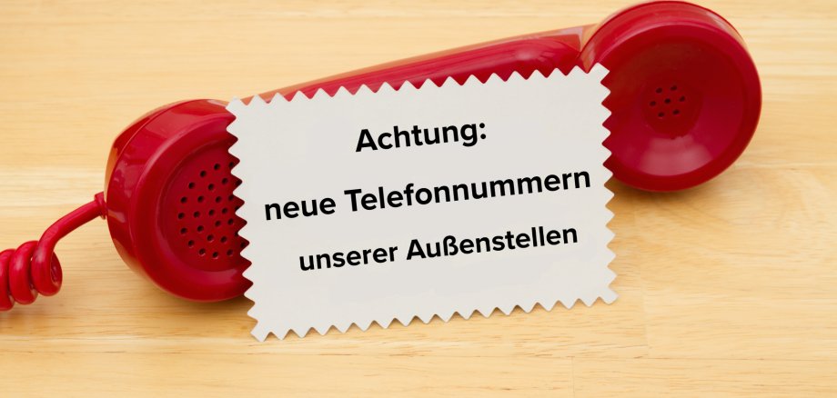 Neue Telefonnummern Roter Telefonhörer mit Telefonschnur. Davor ein weißes Kärtchen mit Hinweistext zu neuen Telefonnummern