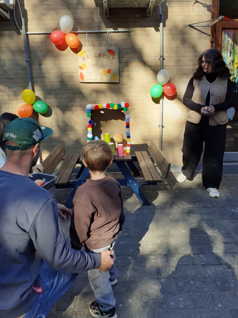 Ein Kind wirft an einer Station mit Ballwerfen einen Ball auf Plastikbecher. Daneben stehen der Vater und eine Erzieherin. 