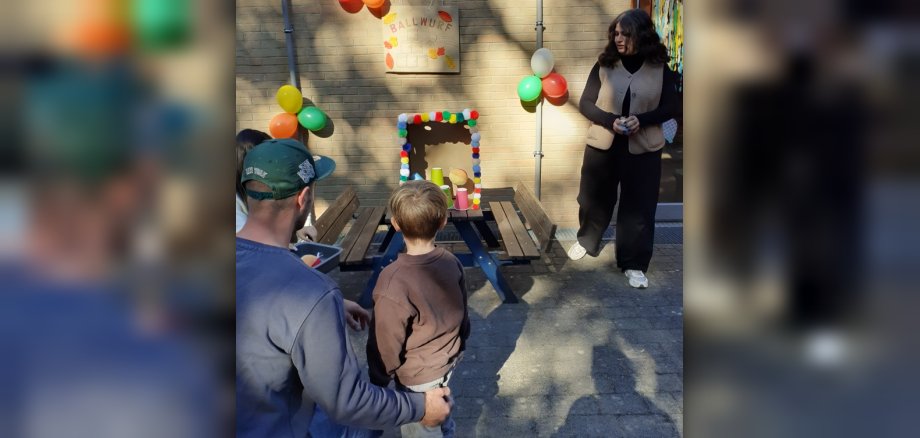 Ballwerfen beim Herbstfest Ein Kind wirft an einer Station mit Ballwerfen einen Ball auf Plastikbecher. Daneben stehen der Vater und eine Erzieherin.