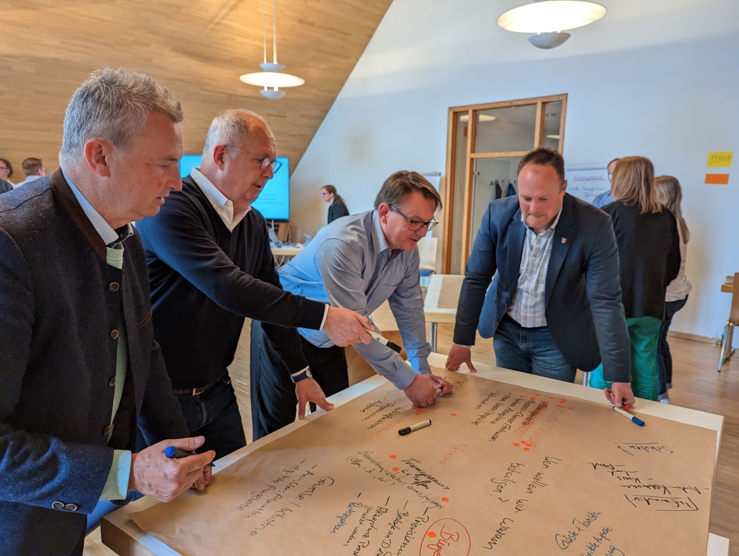 Vier Männer diskutieren an Tisch, auf dem ein Plakat mit Stichwörtern liegt
