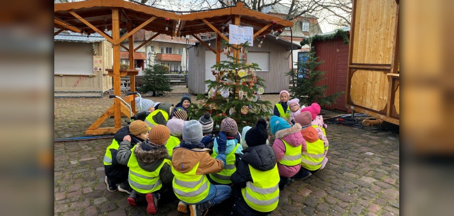 Kita-Kinder sitzen um Weihnachtsbaum auf Marktplatz